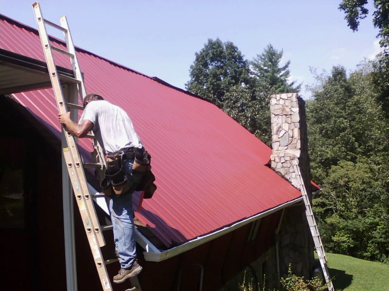 Roofer on a ladder beside a red metal roof and stone chimney — Soffit & Fascia Repair by Roofing Brothers in Buckeye Lake